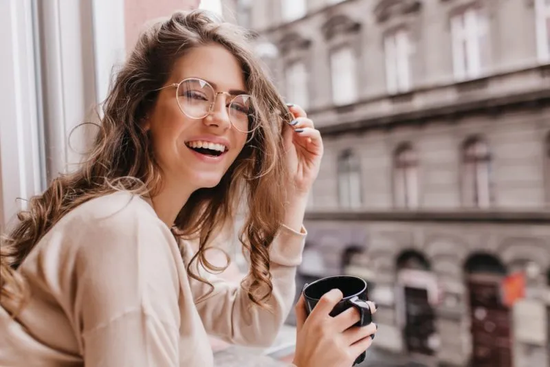 woman in front of window laughing and holding a mug